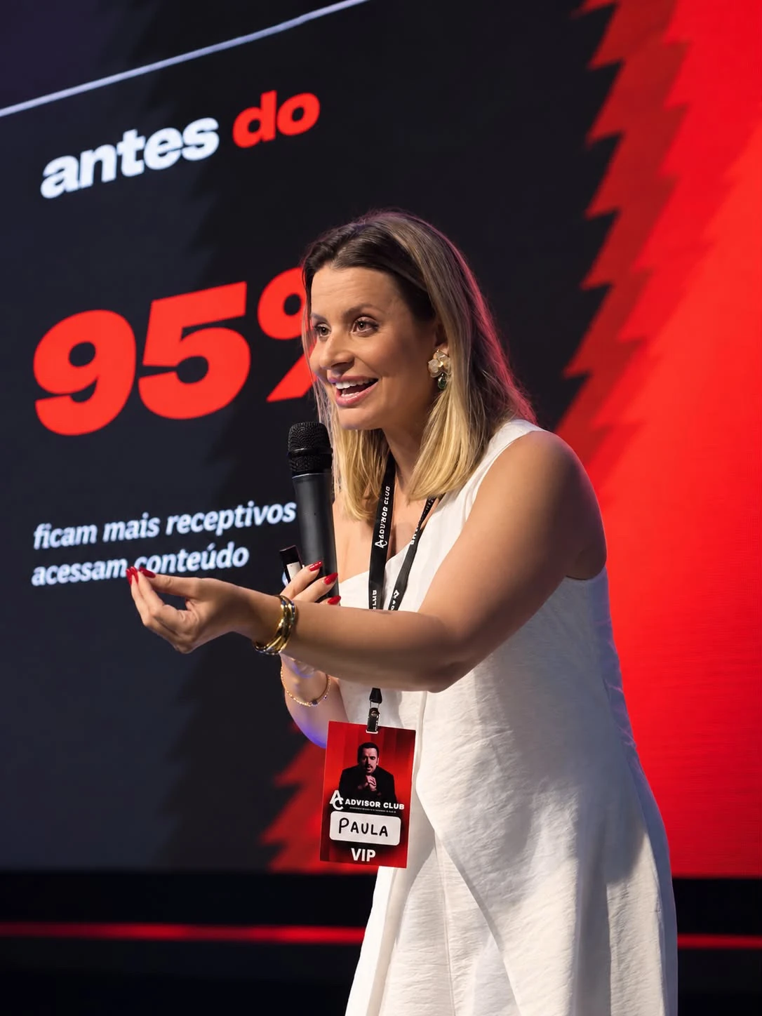 Paula Angelini sorrindo no palco, com o microfone na mão, durante uma palestra