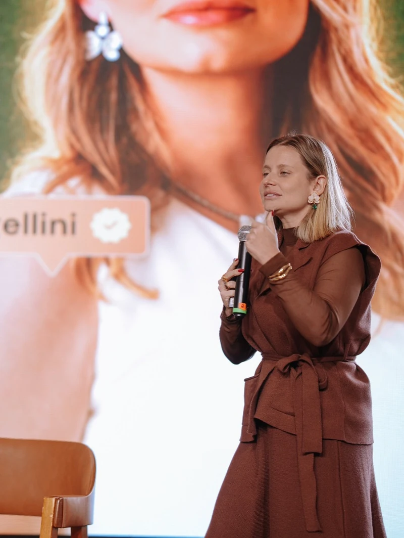 Paula Angelini no palco, com o microfone na mão, diante de um telão com a própria foto